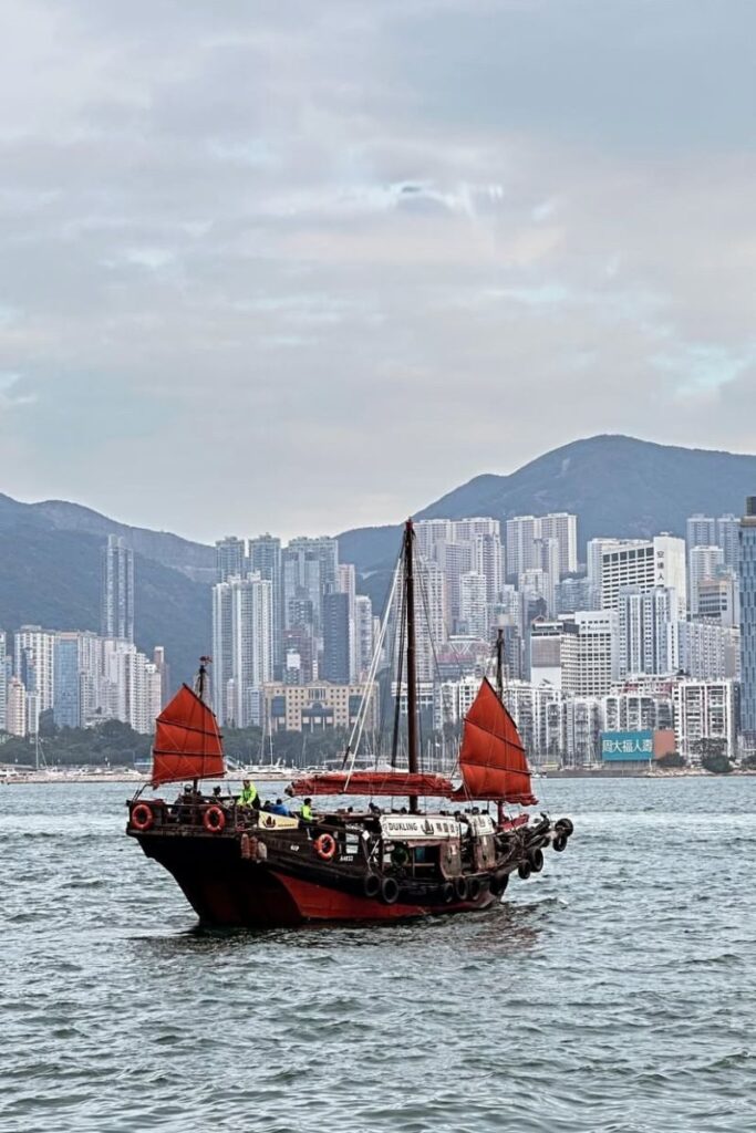 A traditional red-sailed junk boat floats across Victoria Harbour with Hong Kong’s high-rise skyline and mountains in the background. A classic scene when debating Hong Kong vs Singapore.