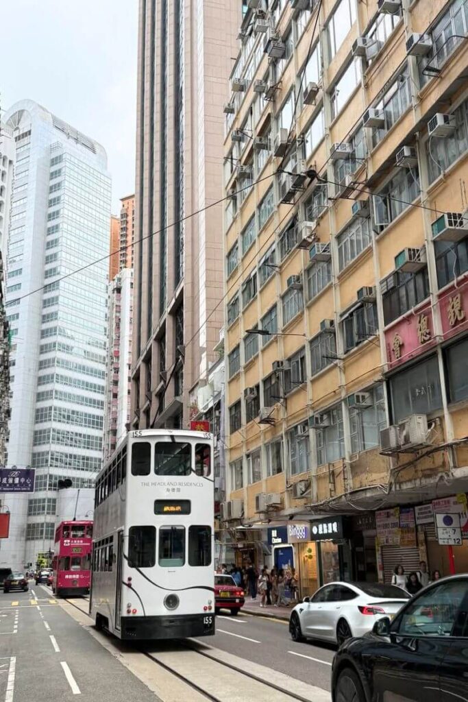 A white double-decker tram passes through a narrow street with tall modern and older buildings in Hong Kong. Riding trams is a must-do and one of the top Hong Kong tips and tricks for affordable city sightseeing.