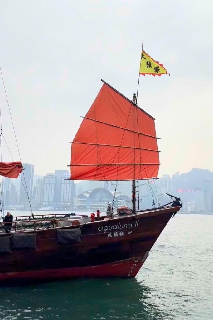 A traditional red-sailed junk boat named “Aqualuna II” sails across Victoria Harbour with Hong Kong skyline behind. Taking a junk boat ride is one of the top travel tips for Hong Kong and a bucket-list experience.