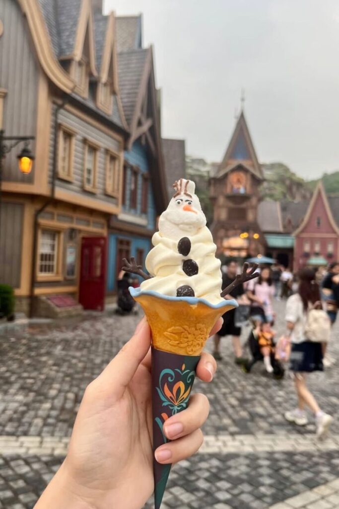 A hand holds an Olaf-themed ice cream cone inside Hong Kong Disneyland with fairytale-style buildings in the background. A fun treat and one of the best tourist tips for Hong Kong disneyland