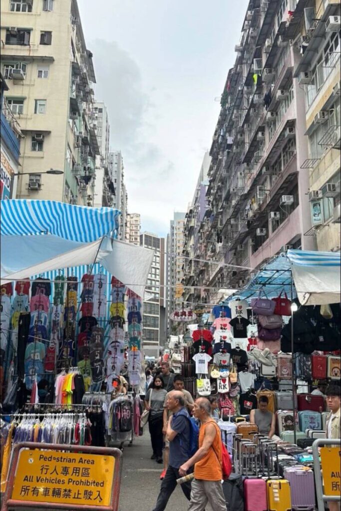 Bustling Hong Kong street market lined with stalls selling clothing, souvenirs, and luggage under tall apartment blocks. A top travel advisory for Hong Kong is to visit night markets for bargains and local culture.