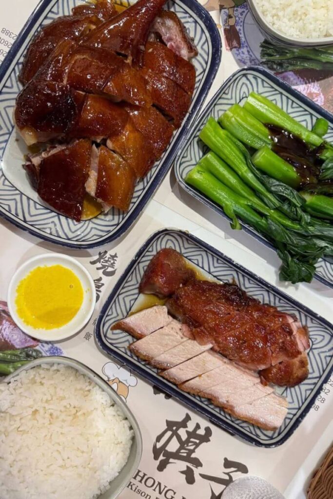 Plates of roast duck, barbecue pork, rice, and steamed greens served at a Hong Kong restaurant. Tasting roast meats is one of the best Hong Kong food tips and an essential thing to know before visiting.