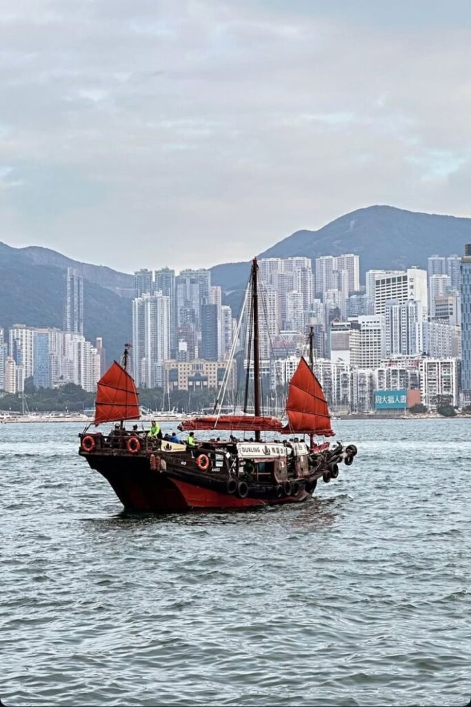 A red-sailed junk boat glides across Victoria Harbour with Hong Kong’s skyline rising behind. Taking a junk ride is a top Hong Kong tourist tip and a must-do cultural experience.