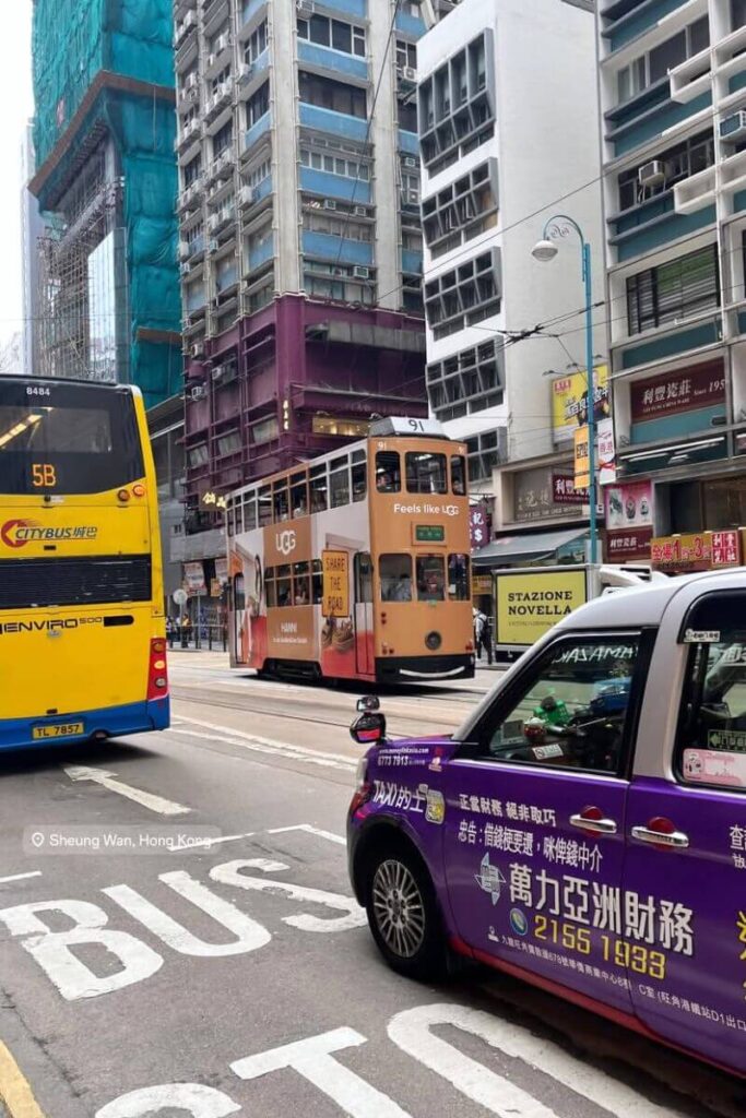 A busy street in Sheung Wan featuring a yellow Citybus, an orange double-decker tram, and a purple taxi driving past old high-rises and local shops. Riding trams through neighborhoods like this is one of the cool things to do in Hong Kong and adds character to any 1 day in Hong Kong experience.