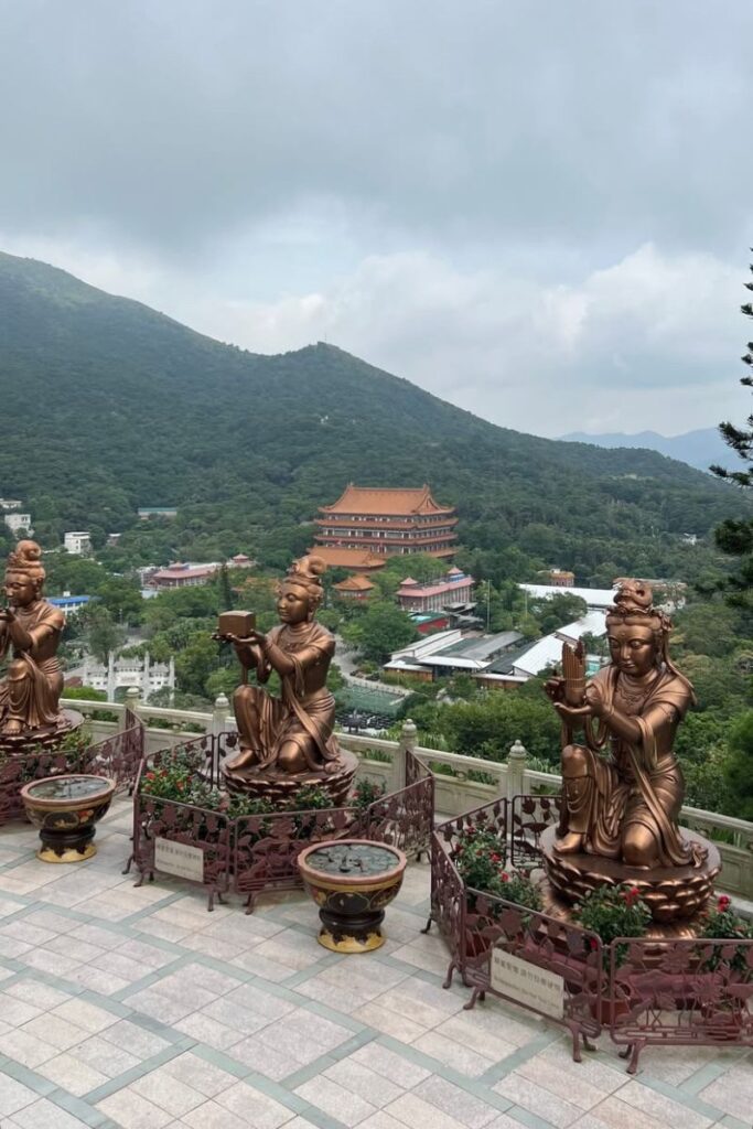 Bronze statues of offering deities at Po Lin Monastery with lush green mountains and a traditional temple in the background. A cultural must-see when considering Hong Kong or Singapore.