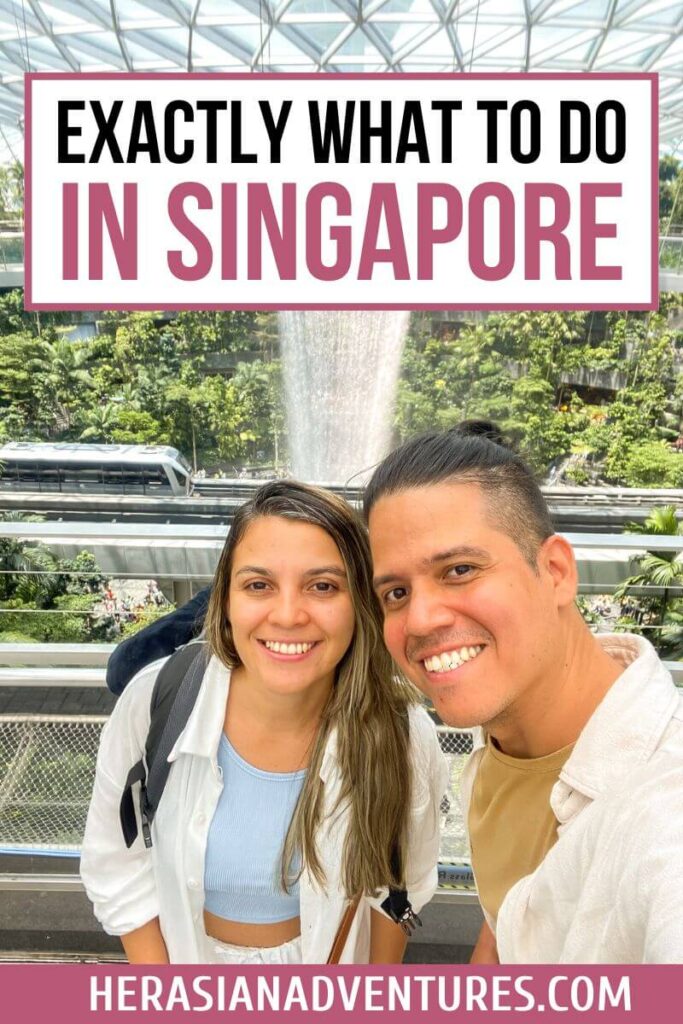 A smiling couple poses in front of the lush Rain Vortex waterfall inside Jewel Changi Airport, under the banner “Exactly What to Do in Singapore.” This stunning indoor attraction is perfect for a layover and ranks high among things to do in Singapore airport.