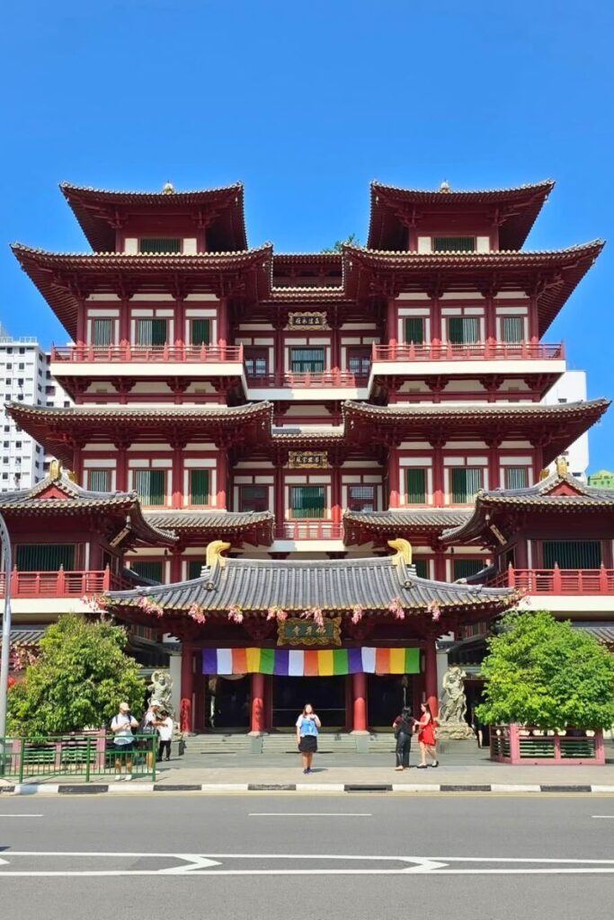 The ornate red-and-white Buddha Tooth Relic Temple in Chinatown stands tall under a clear blue sky, decorated with golden dragons and rainbow prayer flags. This spiritual site is one of the top cultural attractions in Singapore and a peaceful spot to explore heritage.