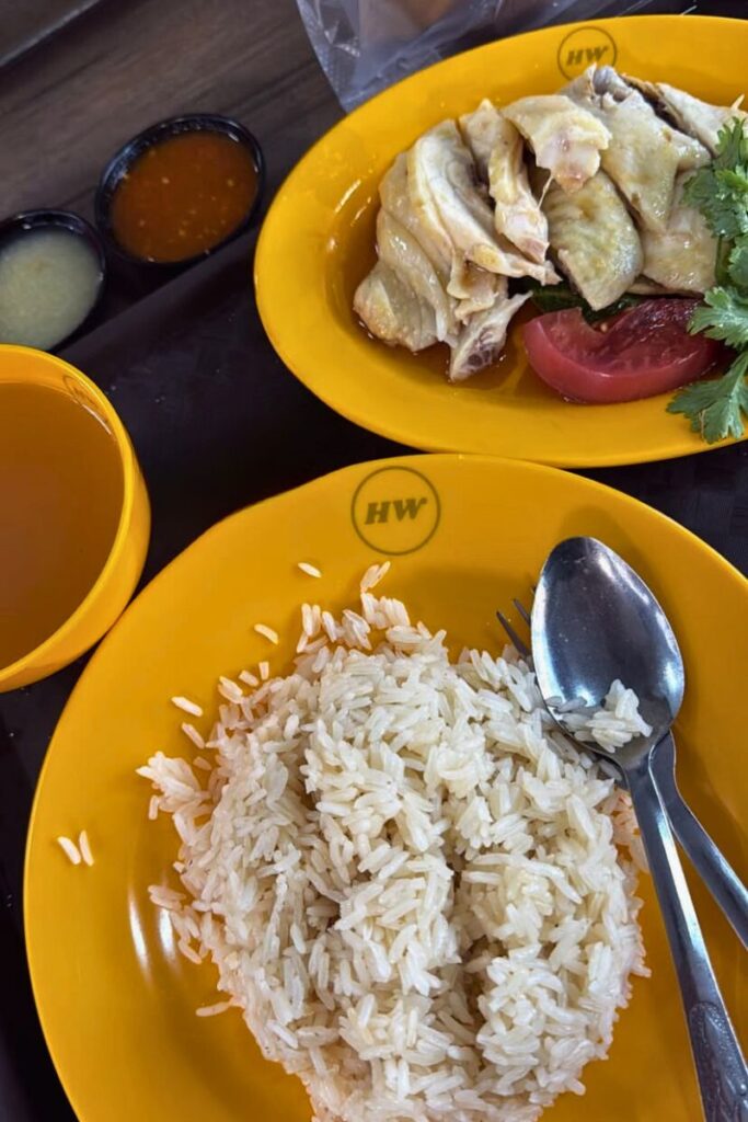 A plate of fluffy white rice served with tender Hainanese chicken, tomato, and coriander, alongside dipping sauces at a local hawker center. Tasting this iconic dish is one of the top foodie things to do in Singapore and a staple of its must-try local cuisine.