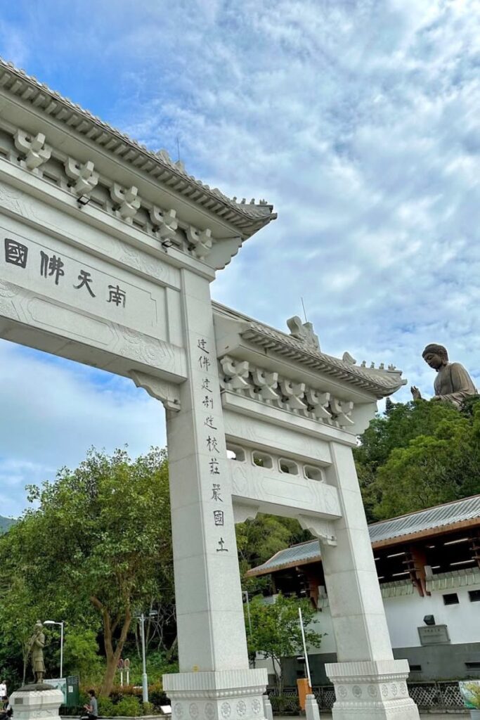 Large white entrance arch with Chinese inscriptions, with the Tian Tian Buddha Hong Kong visible in the background. A must-see landmark on Lantau Island.