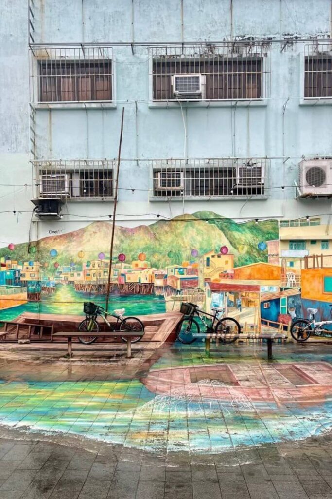 Colorful mural of stilt houses and boats at Tai O Fishing Village, painted against a pale blue wall. This artwork captures the charm of Tai O, often paired with a Lantau Island itinerary to the HK Big Buddha.