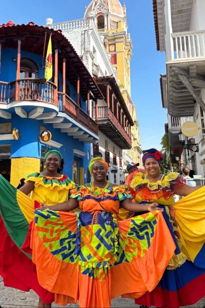 Three women in bright traditional dresses smile in front of colorful colonial buildings in Cartagena, a vibrant choice for those exploring warm countries in December and festive winter travel destinations.