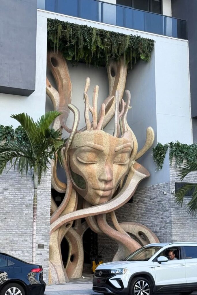 A large artistic sculpture of a serene face decorates a modern building’s entrance lined with palm trees, a creative stop for travelers seeking warm cities in December with unique art scenes.