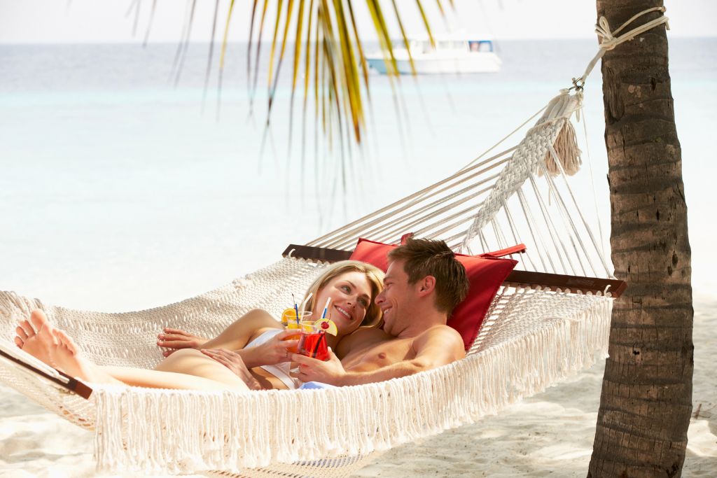 A smiling couple lounges in a hammock under a palm tree with tropical drinks beside a turquoise ocean. This image captures the essence of warm beaches in December and romantic winter getaways in hot countries.
