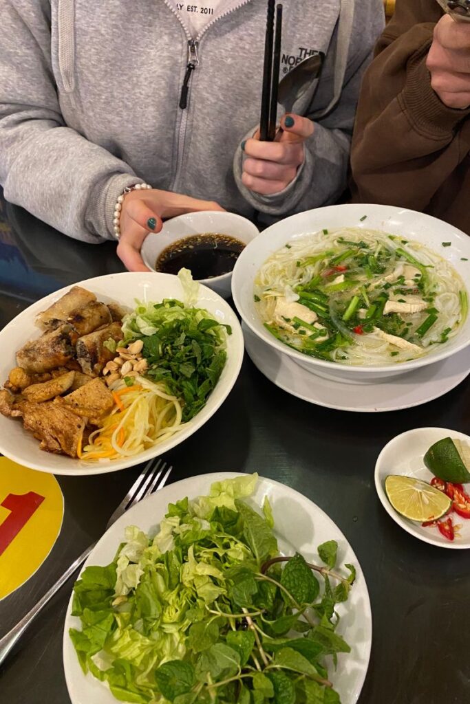 A delicious spread of Vietnamese pho, fried spring rolls, and fresh herbs on a table, shared between two travelers. This cozy setting captures the social side of food hostel culture and is ideal for those discovering local meals while staying in hostels.