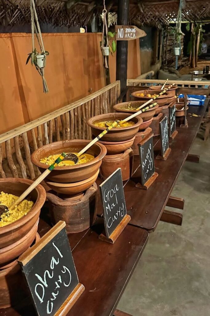 A rustic buffet table lined with clay pots labeled “Dhal Curry” and “Goa Mal Curry,” offering a communal dining experience. This setup embodies hostel kitchen rules and the social dining culture in food hostels.
