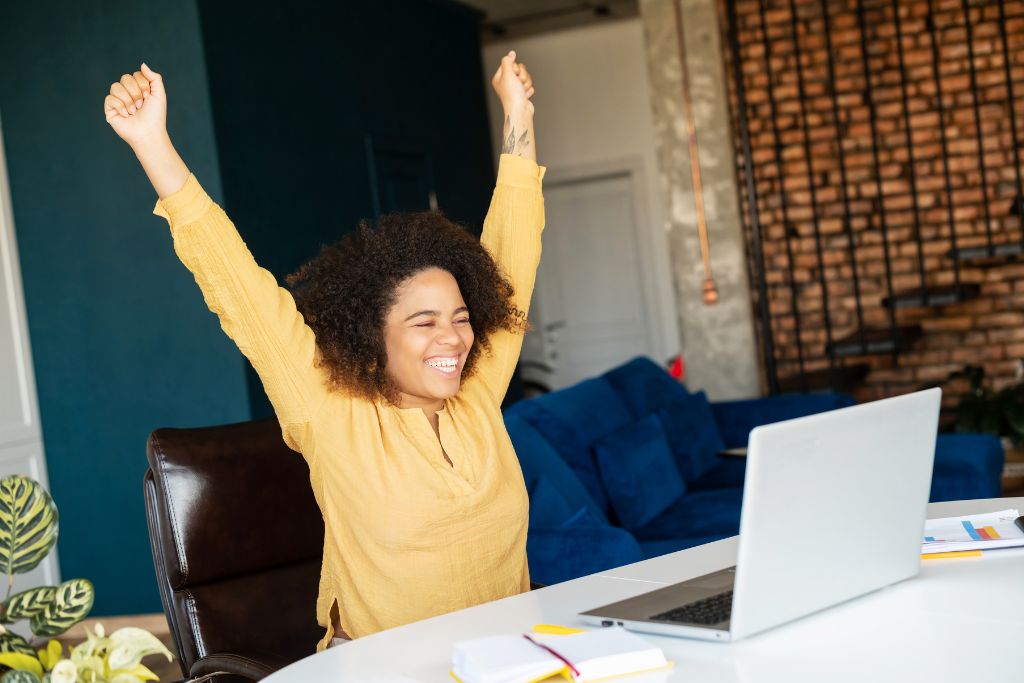 A joyful traveler raises her arms in celebration while sitting at a desk with a laptop. This image conveys the excitement of booking your first hostel stay and discovering hostel tips that make traveling smoother and more affordable.