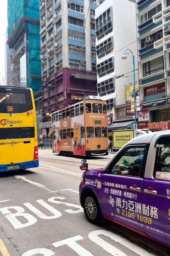 A busy Hong Kong street in Sheung Wan with a double-decker tram, Citybus, and a purple taxi among tall urban buildings. A key travel tip for Hong Kong visitors is to try the tram for an affordable and authentic local experience.