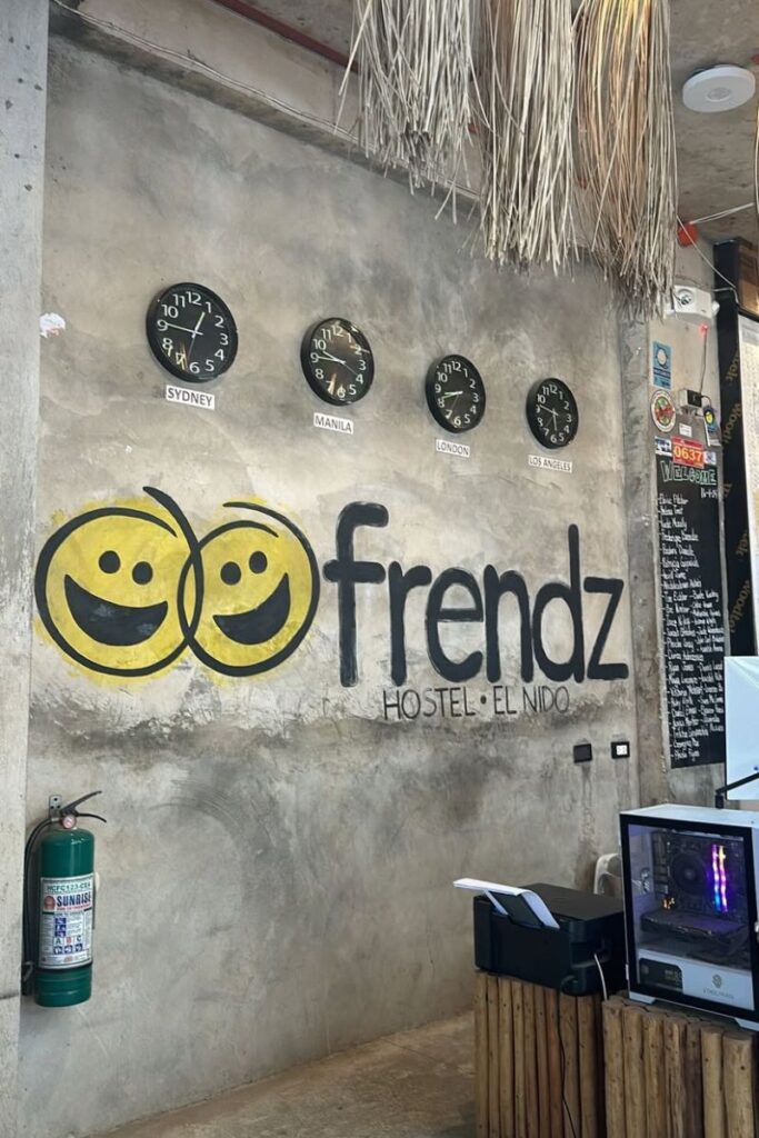 A rustic concrete wall at Frendz Hostel El Nido features clocks showing world times and a cheerful yellow logo. This photo highlights hostel standards and a welcoming atmosphere for travelers staying at a hostel abroad.