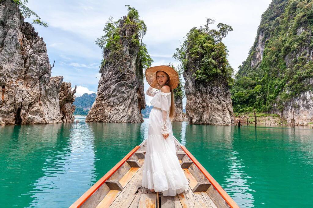 A woman in a white dress and straw hat poses on a wooden boat surrounded by dramatic limestone cliffs and emerald water. Though not a hostel image, it represents travel adventures that pair well with budget-friendly hostel stays and Southeast Asia travel tips.