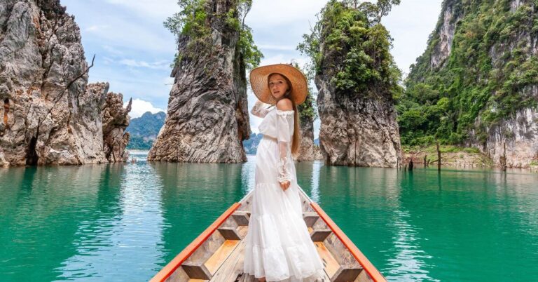 A woman in a white dress and wide sunhat stands gracefully on a wooden boat surrounded by towering limestone cliffs and emerald-green water. This stunning tropical scene highlights one of Asia’s top warm destinations in October, ideal for dreamy winter vacations and travelers seeking hot countries in December.