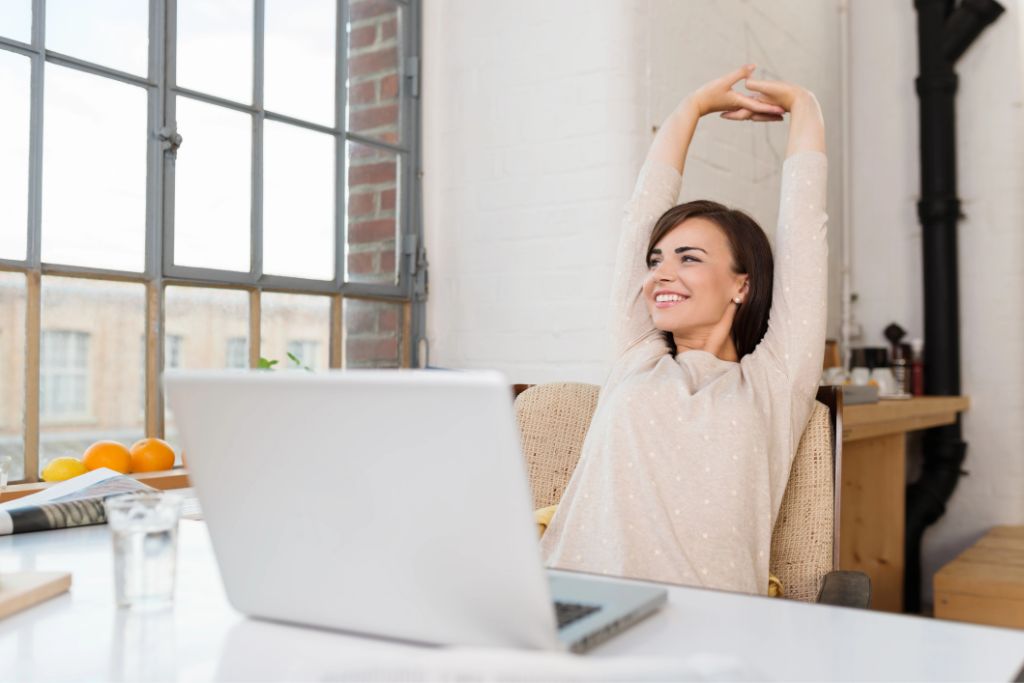 A relaxed woman stretches in her home office beside a laptop, suggesting work-from-home habits that help you save money for travel and reduce daily expenses for a holiday fund.