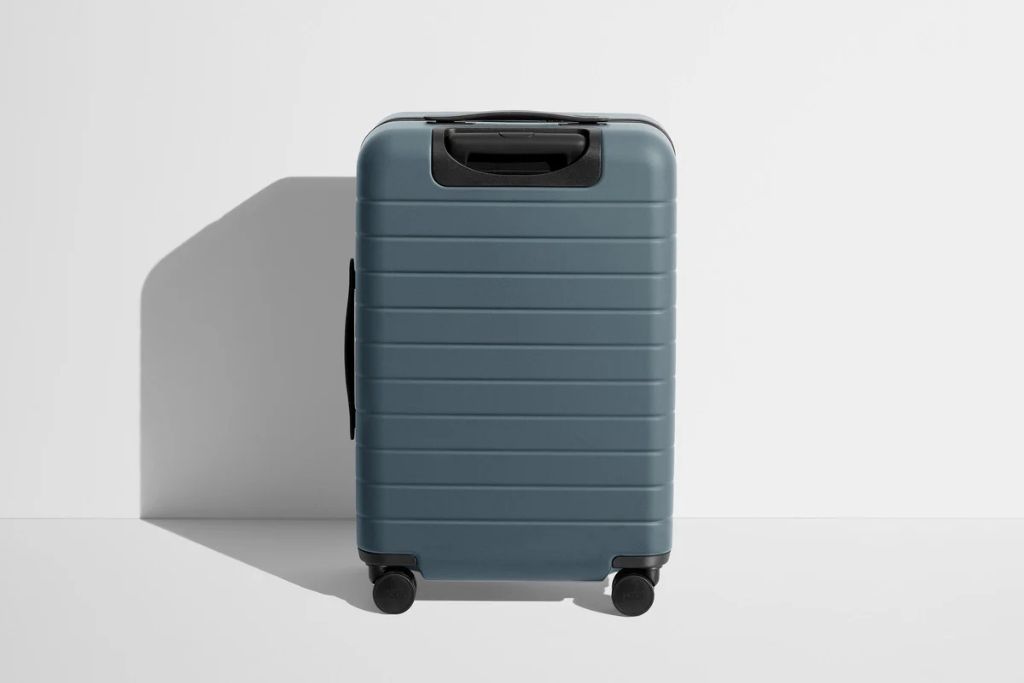A minimalist grey suitcase stands upright against a white wall, symbolizing travel fund money box goals and how much money you need to travel when planning your next trip.