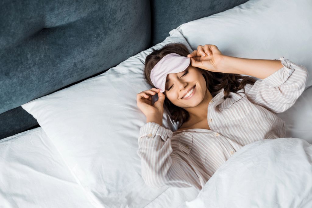 A woman rests in bed with an eye mask, highlighting simple lifestyle swaps—like staying in—to save money for a vacation and build travel savings faster.