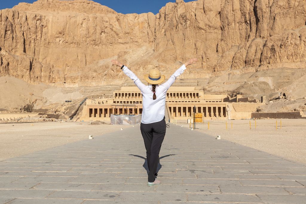A traveler raises her arms toward an ancient stone temple, emphasizing how much money you need to travel and the reward of saving for a holiday.