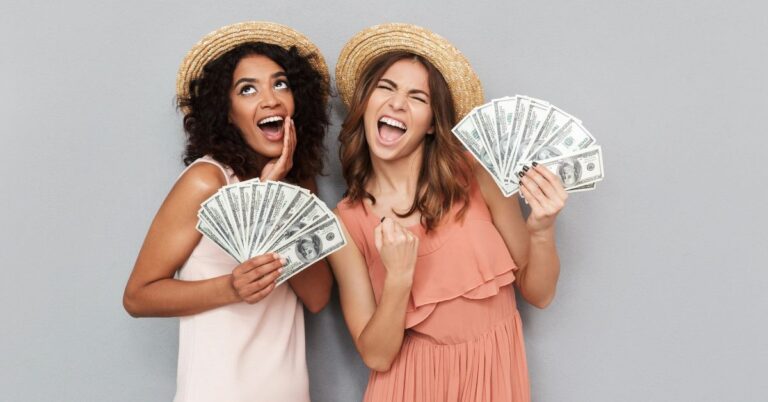 Two excited women in summer hats hold fanfuls of dollar bills, showing how to save money for travel and build travel savings for an affordable holiday.