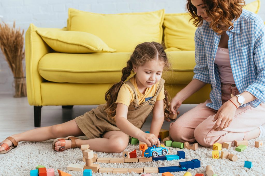 A young girl builds a toy track on the floor with her nanny, suggesting babysitting is a great way to save money for travel as a student and simple habits that support travel savings goals.