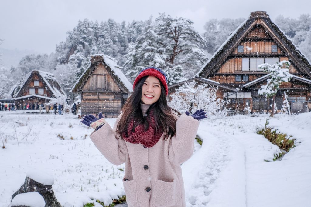 A woman smiles in a snowy village with traditional wooden houses, offering cozy winter imagery ideal for a Japan travel vision board or aesthetic travel vision board wallpaper.