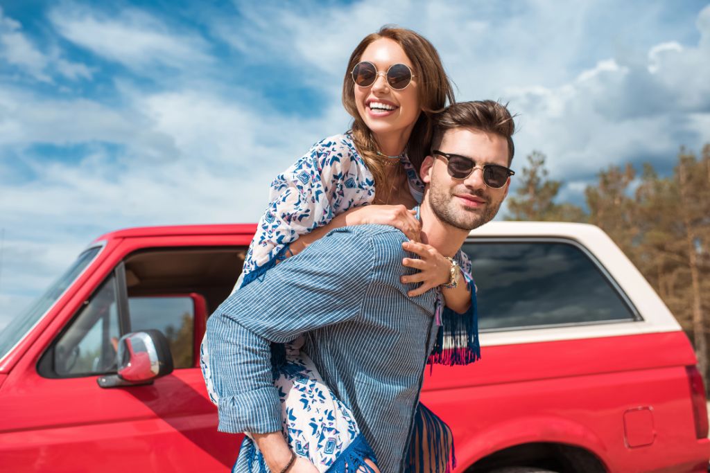 A smiling couple poses in front of a red car during a sunny road trip, capturing joyful moments ideal for a travel with family vision board and dream travel vision board pictures.