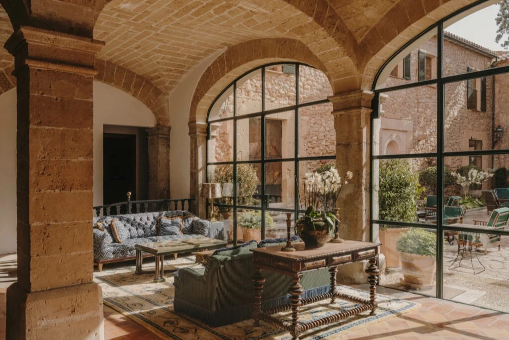 Elegant sofas sit beneath vaulted stone arches beside tall windows overlooking a peaceful courtyard garden. This character-filled setting suits travelers searching where to stay in Mallorca for first time visits or a relaxing Mallorca base.