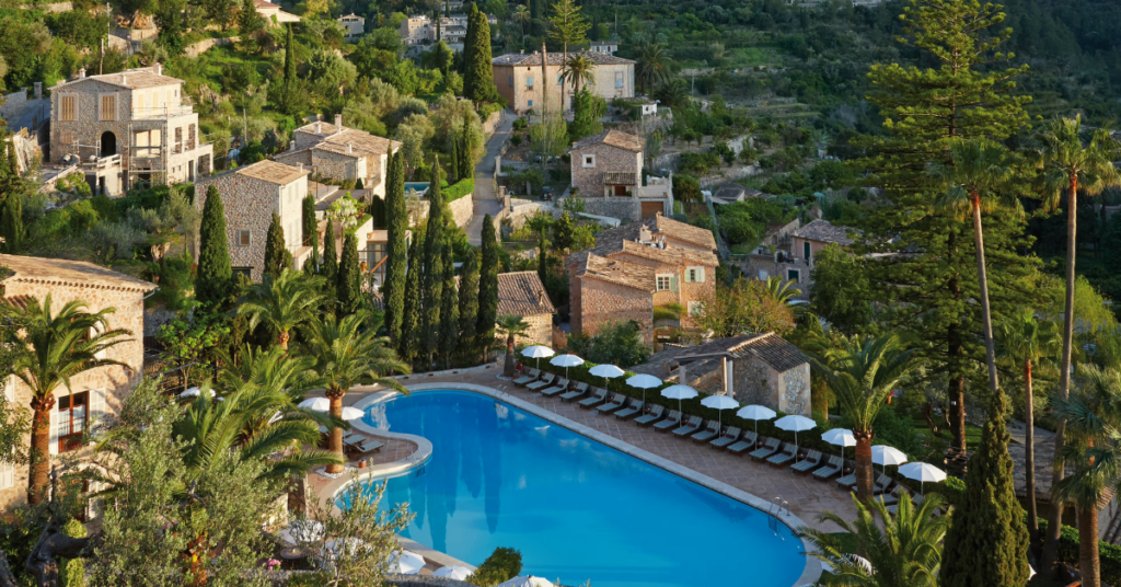 A large blue pool sits among palm trees, cypress-lined stone villas, and lush mountain scenery in the Tramuntana foothills. A dream setting for where to stay in Mallorca for couples, cycling holidays, or a luxury countryside escape.